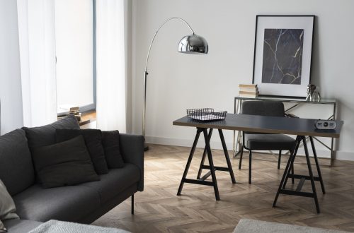 kadr przedstawiający fragment wnętrza mieszkania z sofą, biurkiem, lampą stojąca i podłogą z paneli winylowych ułożonych w jodełkę